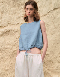 Woman wearing a light blue sleeveless top and white pants against a beige stone wall.