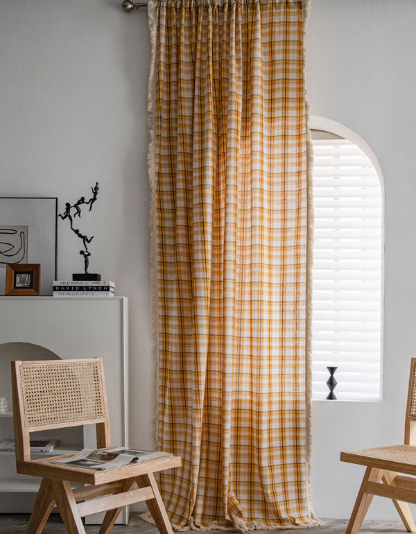 A set of yellow and blue checkered cotton curtains with a tassel edge hanging on a window in a room with a white wall and a wooden chair.