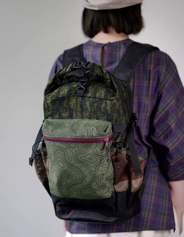Compact terrain‑print roll top backpack worn cross‑body, showing front zip pocket and side mesh pockets.