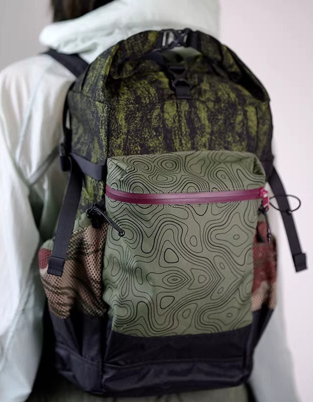 Green and black roll top backpack with topographic print front pocket and side mesh pockets resting in car trunk.
