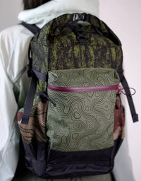 Green and black roll top backpack with topographic print front pocket and side mesh pockets resting in car trunk.