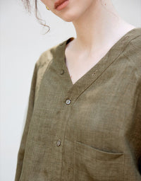 Olive green linen V‑neck blouse with button front, chest pocket and elastic hem, worn with high‑waisted wide‑leg pants.