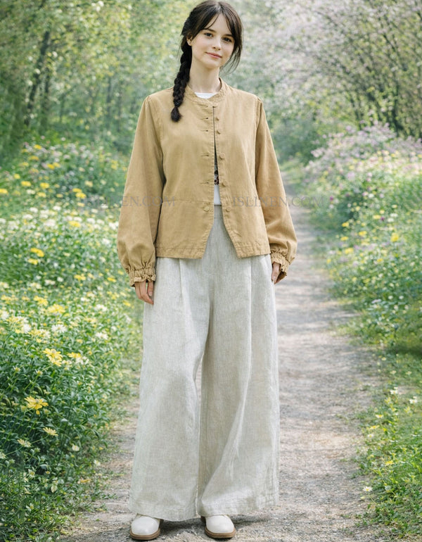 Woman in a beige jacket and light pants standing on a path surrounded by greenery and flowers.