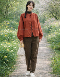 Woman in a rust-colored jacket and brown pants standing on a path with flowers and trees in the background
