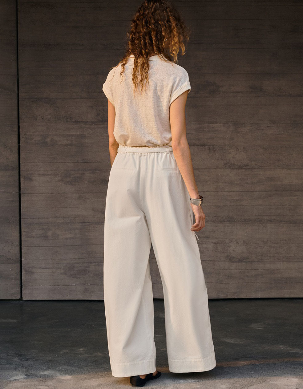 Woman wearing a beige top and wide-leg pants against a dark wooden wall.