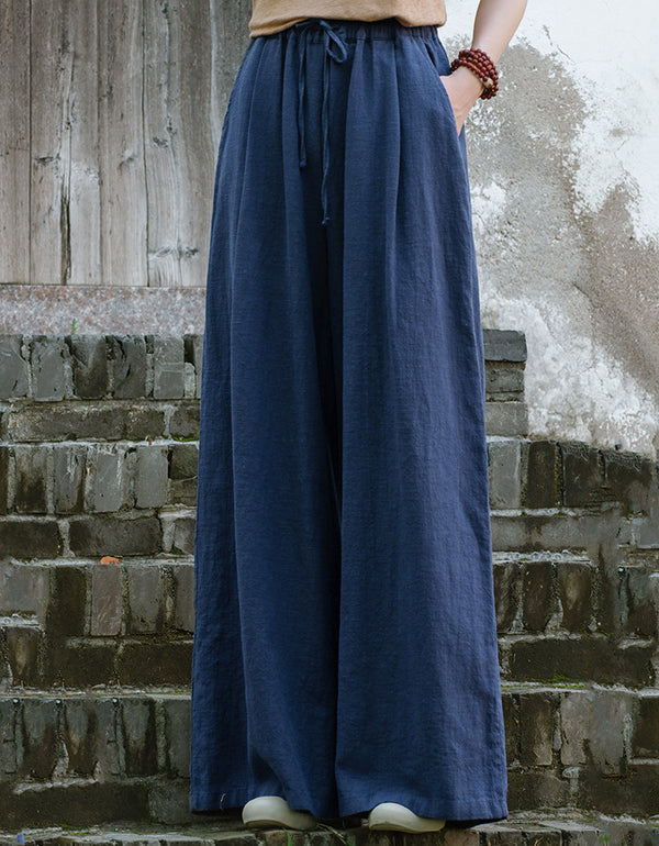 A pair of high-waisted, wide-leg blue linen pants with an elastic drawstring waist, worn by a model standing in front of a rustic background.