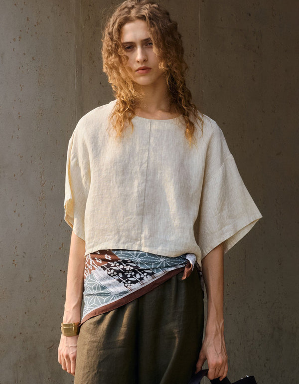 Natural beige boxy linen short sleeve top with wide sleeves and cropped length, worn with dark pants and printed scarf at waist.
