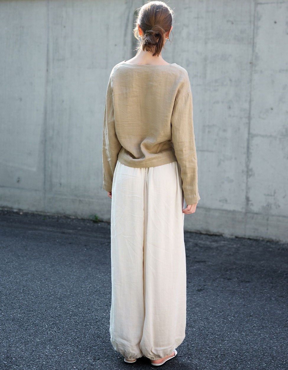 Person wearing a beige blouse and white pants standing against a concrete wall.