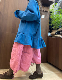 Woman modeling a relaxed blue ruffle-hem shirt with pink trousers in a warm indoor setting.