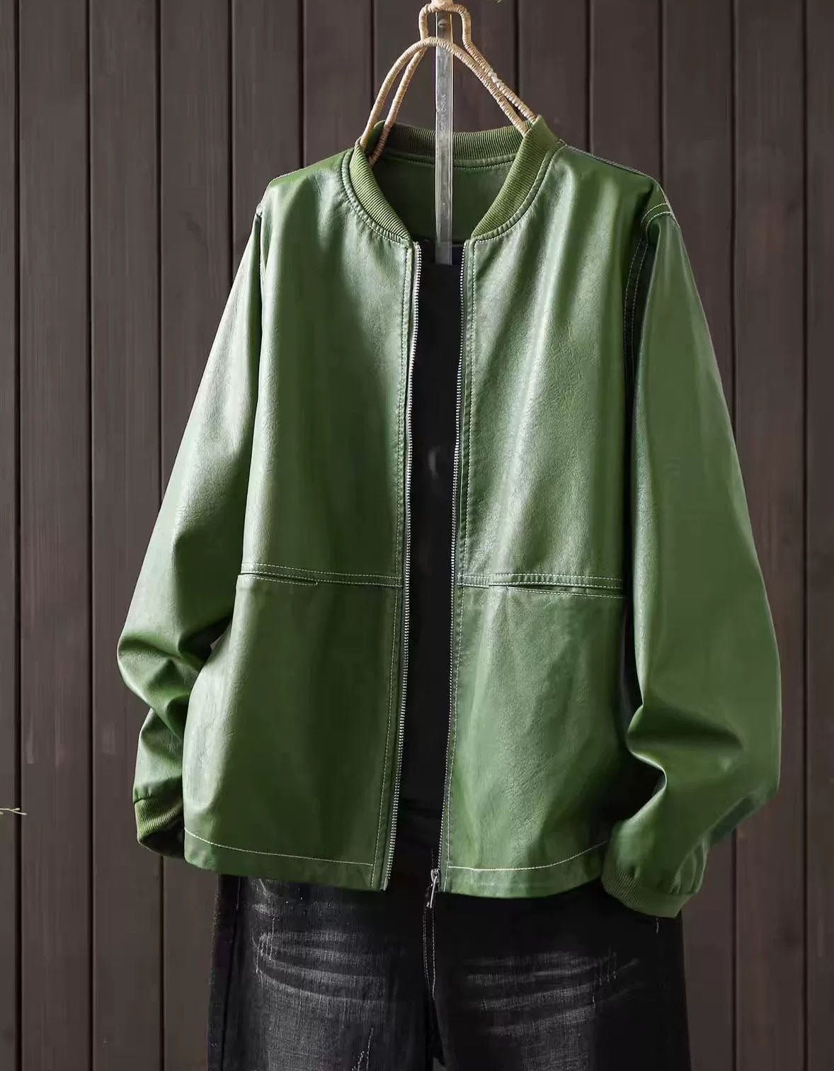 Green faux leather bomber jacket with zip-up front hanging against a dark wood panel background