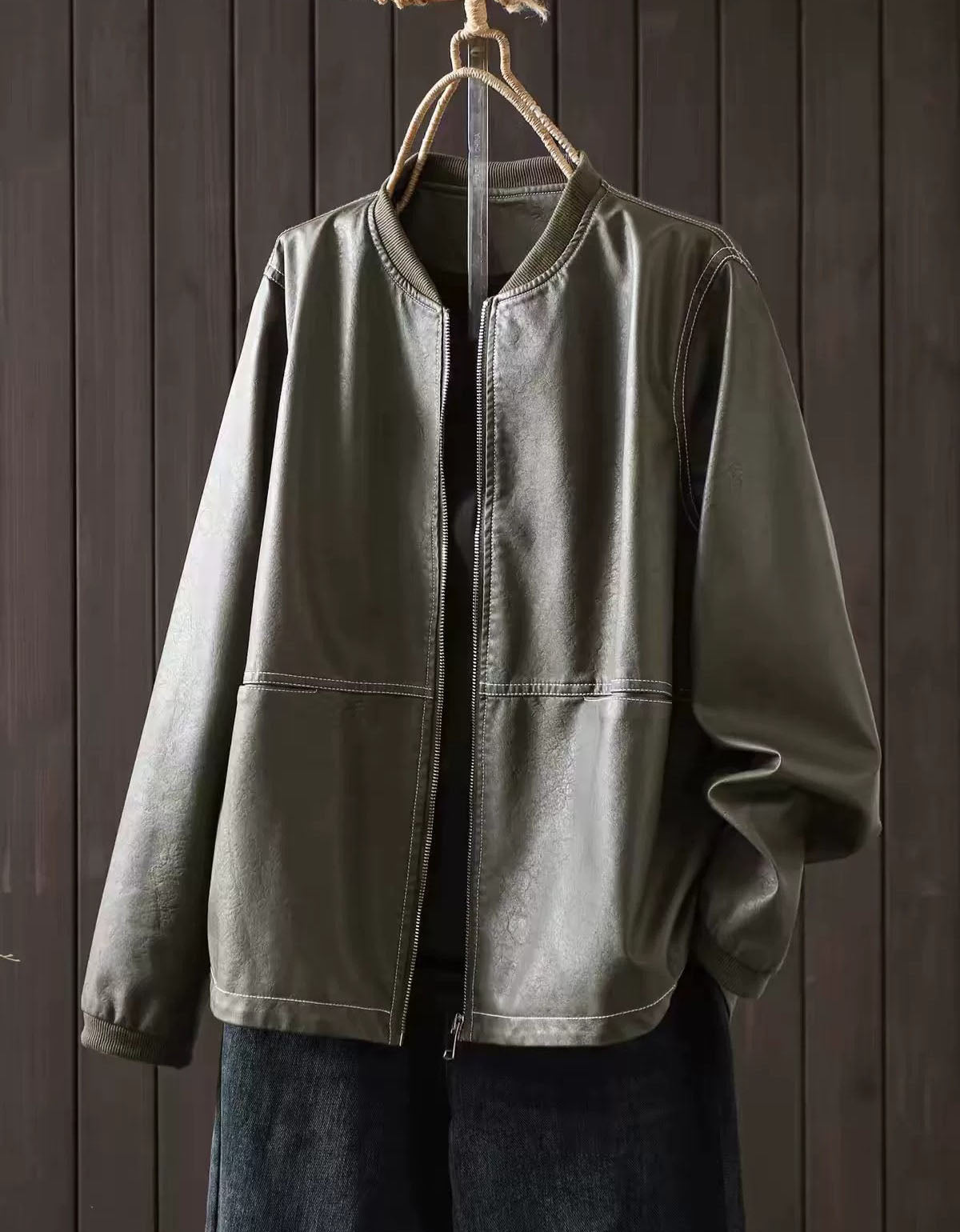 Olive green faux leather bomber jacket with zip-up front hanging against a dark wood panel background