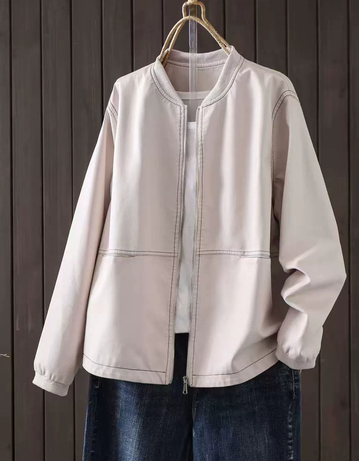 Beige faux leather bomber jacket with zip-up front hanging against a dark wood panel background