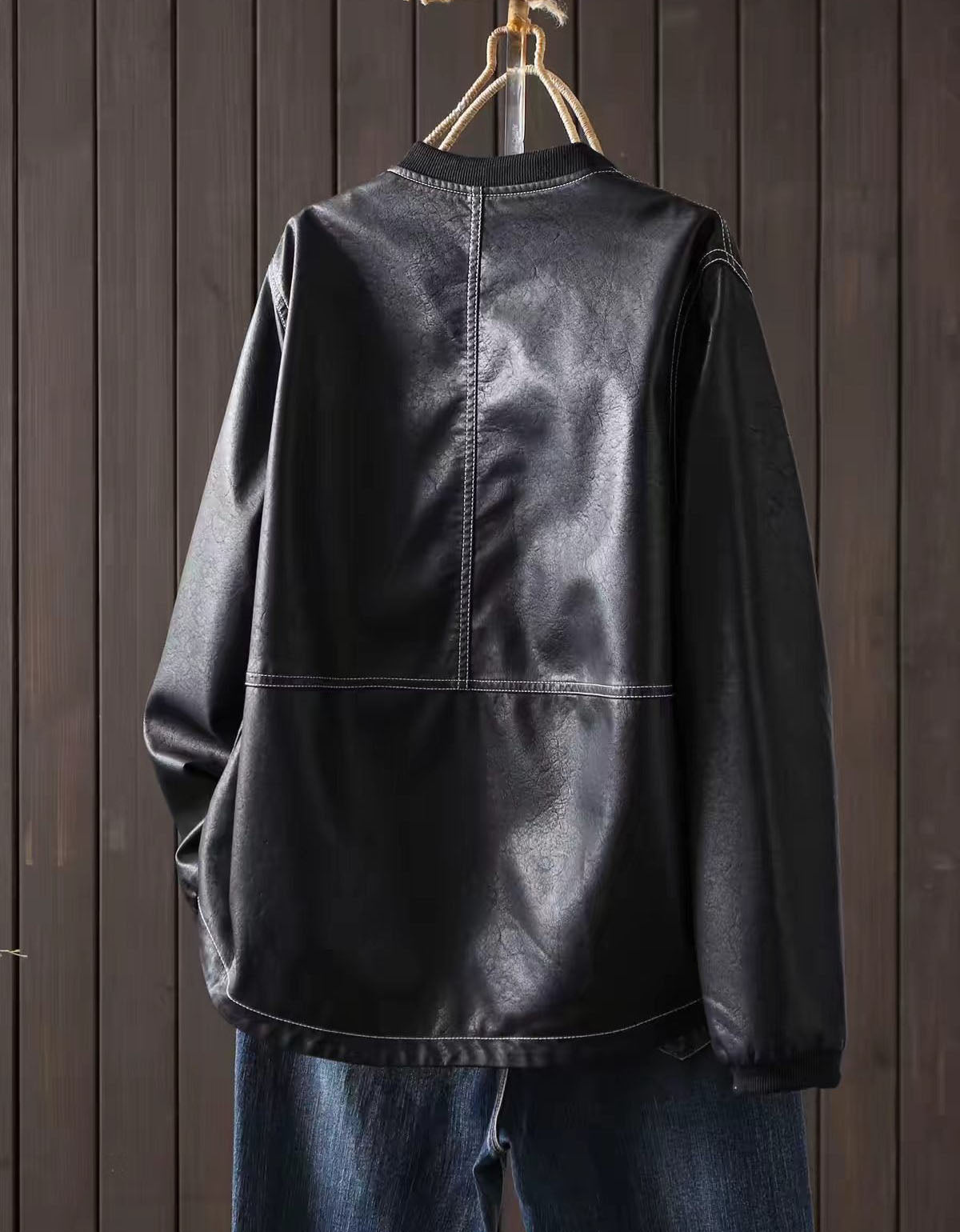 Black faux leather bomber jacket with zip-up front hanging against a dark wood panel background