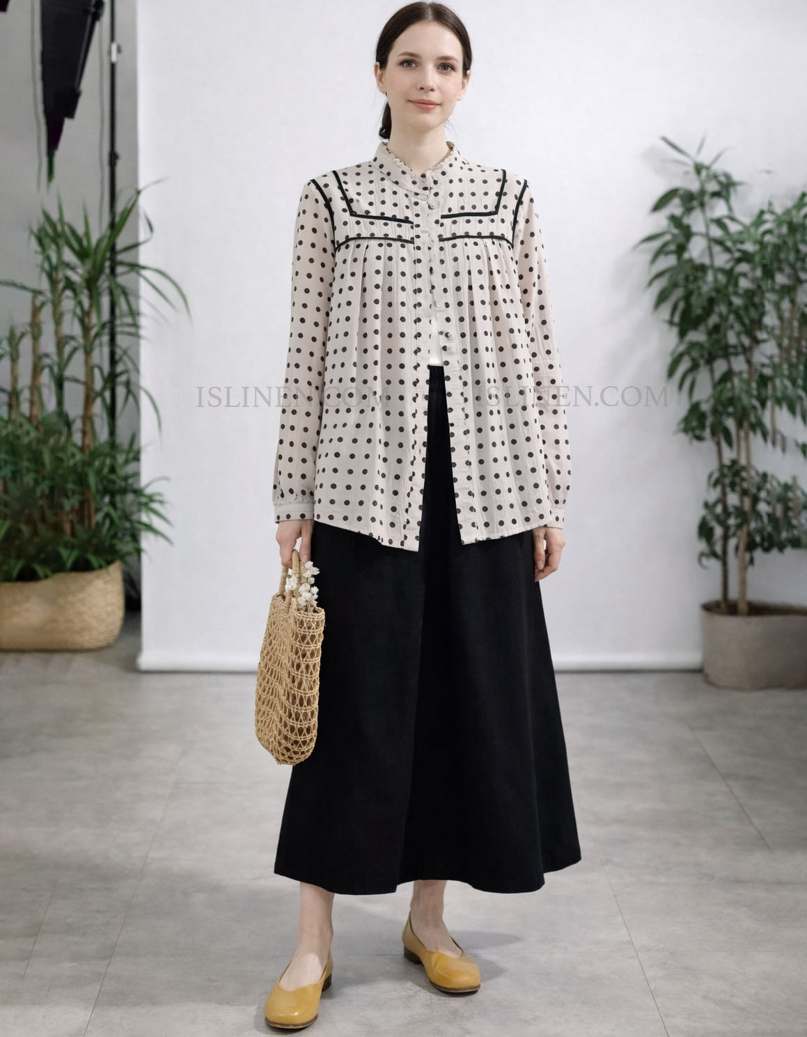 Woman wearing a patterned blouse and black skirt with yellow shoes, standing in a minimalistic room with plants.