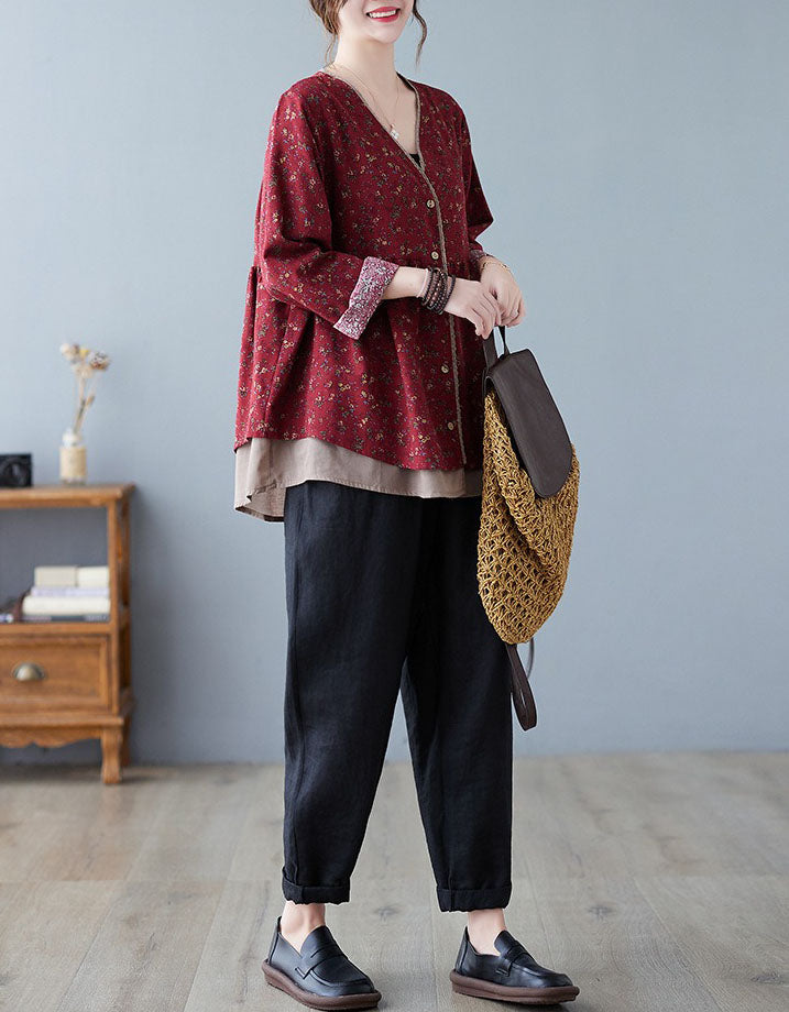Woman wearing a red patterned top and black pants, holding a brown bag against a gray wall.