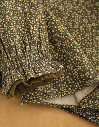 Close-up of a dark olive green fabric with a dense small floral print in cream and yellow, featuring a gathered ruffled sleeve and a solid tan surface underneath.